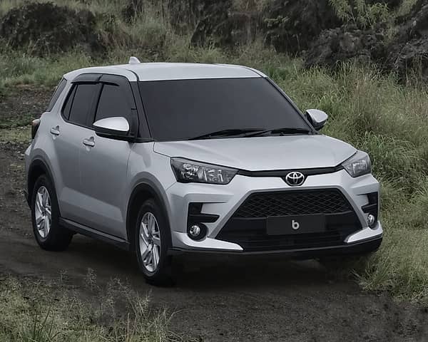 Silver Toyota Raize SUV driving on a dirt road in a grassy, hilly landscape.