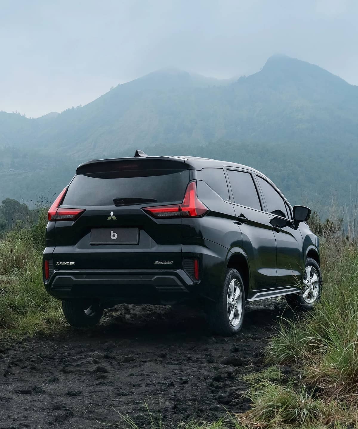 Black Mitsubishi Xpander parked on a dirt road in a rural Balinese landscape with rolling hills and lush vegetation.